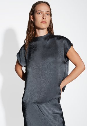Schwarze, strukturierte Bluse mit kurzen Ärmeln und rundem Ausschnitt. Das Material hat einen leichten Glanz und fällt locker über den Körper.