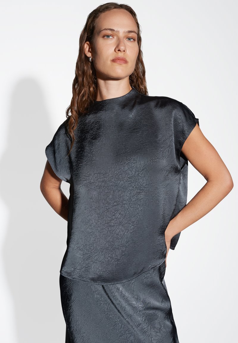 Schwarze, strukturierte Bluse mit kurzen Ärmeln und rundem Ausschnitt. Das Material hat einen leichten Glanz und fällt locker über den Körper.