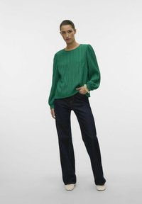 Grønne rillede langærmede top med rund halsudskæring og pufærmer, parret med mørke brede jeans og lyse sko.