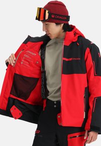 Rote und schwarze Spyder-Ski-Jacke aus wasserdichtem Material, mit Reißverschlusstaschen, verstellbarer Kapuze und Mesh-Innenfächern.