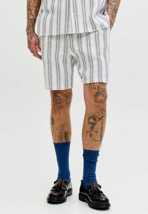 Mężczyzna ubrany w białe szorty i pasującą koszulę z pionowymi czarnymi paskami, niebieskie skarpetki, czarne mokasyny z frędzlami oraz widoczne tatuaże na ramionach i nogach.