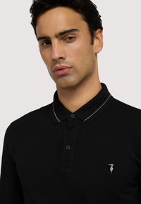 Czarna koszulka polo z długim rękawem i listwą na dwa guziki, z subtelnym białym paskowanym kołnierzykiem oraz małym logo na lewej piersi.