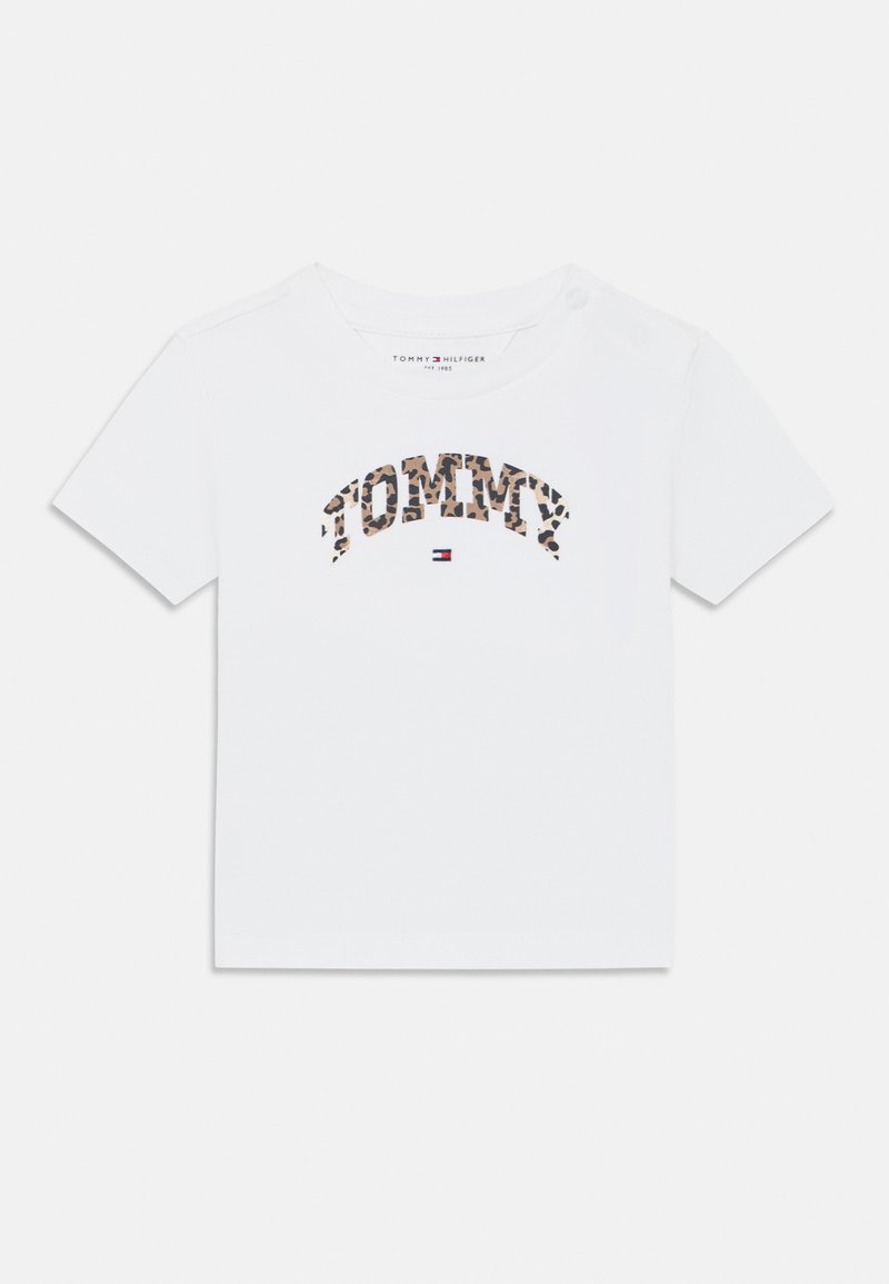 White short-sleeve t-shirt with "TOMMY" in leopard print lettering. Features a round neck and button detail on one shoulder. Smooth cotton fabric.