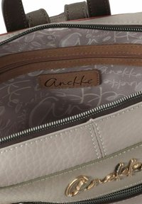 Beige Innenseite einer Handtasche mit glatter Textur, markiert mit "Anekke", ausgestattet mit einer Reißverschlusstasche und einem sich wiederholenden hellen Muster.