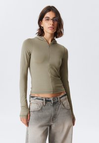 Olive green cropped hoodie with long sleeves, zip front closure, and smooth texture, paired with light grey faded jeans.