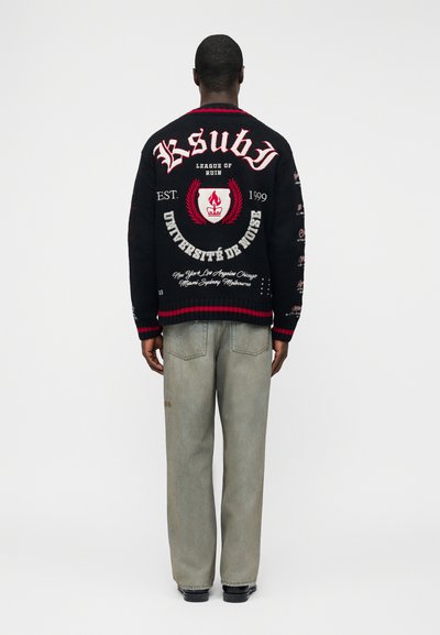 Uomo in piedi di spalle che indossa un maglione nero con testo ed emblema rosso e bianco, abbinato a jeans grigi larghi e scarpe nere.