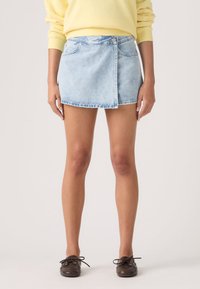 Γυναίκα που φοράει μίνι φούστα από ανοιχτό μπλε τζιν, κίτρινο φούτερ και καφέ παπούτσια με κορδόνια, στέκεται μπροστά σε απλό φόντο.