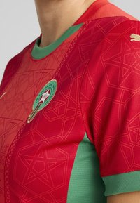 Czerwona koszulka sportowa z geometrycznymi wzorami gwiazd, zielonym kołnierzykiem i bocznymi panelami oraz zielonym emblematem z arabskim napisem i koroną.