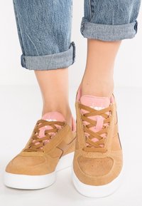 Hnědé semišové tenisky s růžovými akcenty, kulatou špičkou, perforovanými detaily a rovnou bílou podrážkou. Šňůrky jsou hnědé a jazyk je růžový.