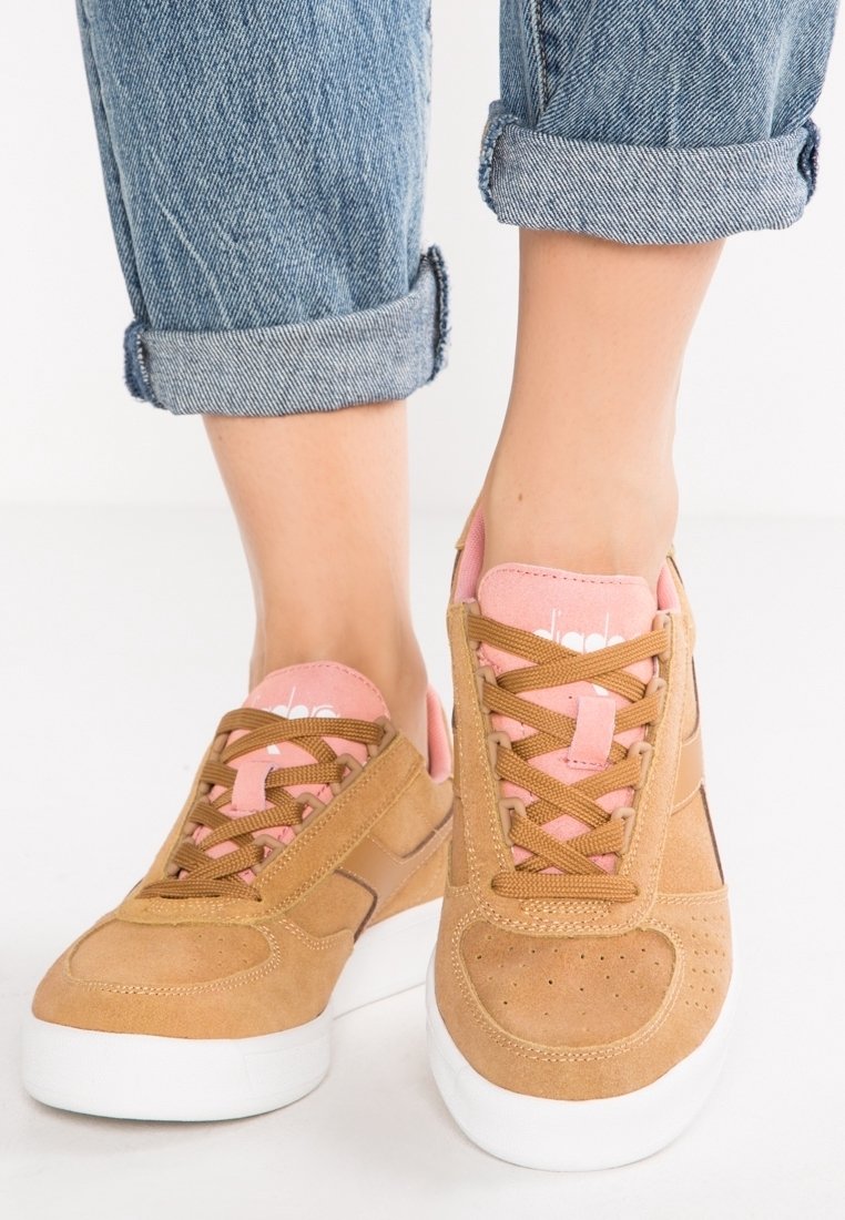 Hnědé semišové tenisky s růžovými akcenty, kulatou špičkou, perforovanými detaily a rovnou bílou podrážkou. Šňůrky jsou hnědé a jazyk je růžový.