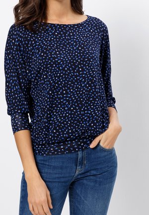 Femme portant un haut à manches longues bleu marine avec des taches bleues et blanches dispersées, associé à un jean en denim bleu, main dans la poche.
