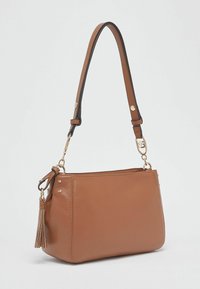 Sac à bandoulière en cuir synthétique marron avec une bandoulière amovible, des finitions dorées et un pompon latéral. Caractérisé par une forme rectangulaire structurée.