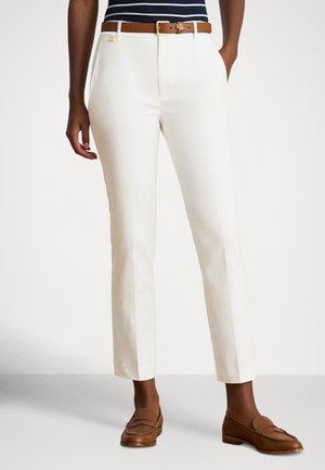 Persoon die witte slim-fit broeken, een bruine leren riem en bruine loafers draagt, staand tegen een effen lichte achtergrond.
