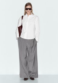 Femme portant une chemise blanche surdimensionnée, un pantalon large gris, des chaussures foncées, des lunettes de soleil sombres, des boucles d'oreilles créoles et portant un grand sac à bandoulière brun.