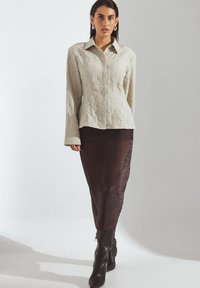 Lichtbeige, gestructureerd overhemd met lange mouwen, gecombineerd met een donkerbruine, strakke rok en zwarte kniehoge laarzen.