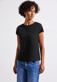 T-shirt noir à manches courtes avec un col rond, doté de clous métalliques sur les épaules et d'une coupe décontractée. À porter avec un jean en denim bleu.