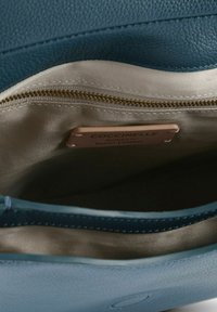 Interior de bolso de cuero azul con forro beige claro, un bolsillo con cremallera y una etiqueta que dice "Coccinelle Cuero Italiano Auténtico".