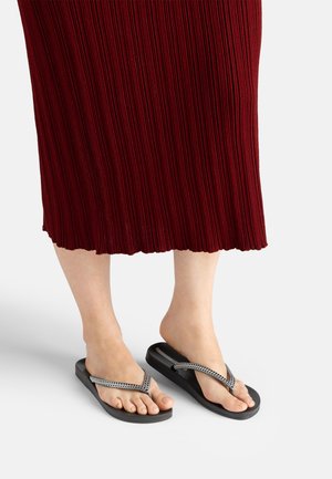 Barfuß mit schwarzen Flip-Flops mit geflochtenen Riemen, die nebeneinander stehen, teilweise bedeckt von einem burgunderfarbenen Faltenrock.