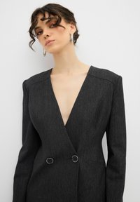 Donkergrijze op maat gemaakte blazer met een diepe V-hals, gestructureerde schouders en dubbele ronde knoopaccenten. Glad, textuurstof.