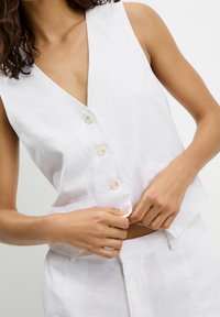 Haut sans manches en lin blanc avec un col en V profond, orné de quatre boutons et d'une texture subtile. Porté avec un pantalon assorti de couleur claire.