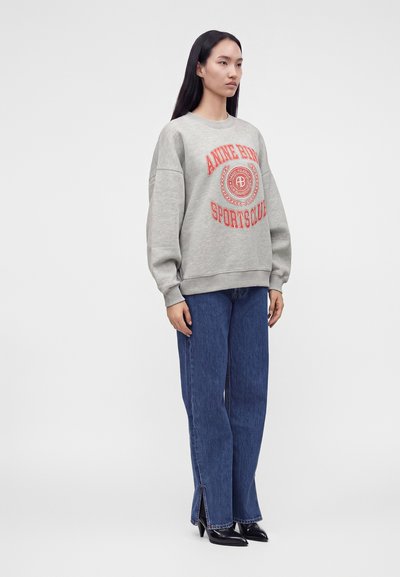 Femme aux longs cheveux foncés porte un sweat-shirt gris oversize avec un texte rouge, un jean bleu à jambes larges avec des fentes à l'avant et des chaussures noires pointues.