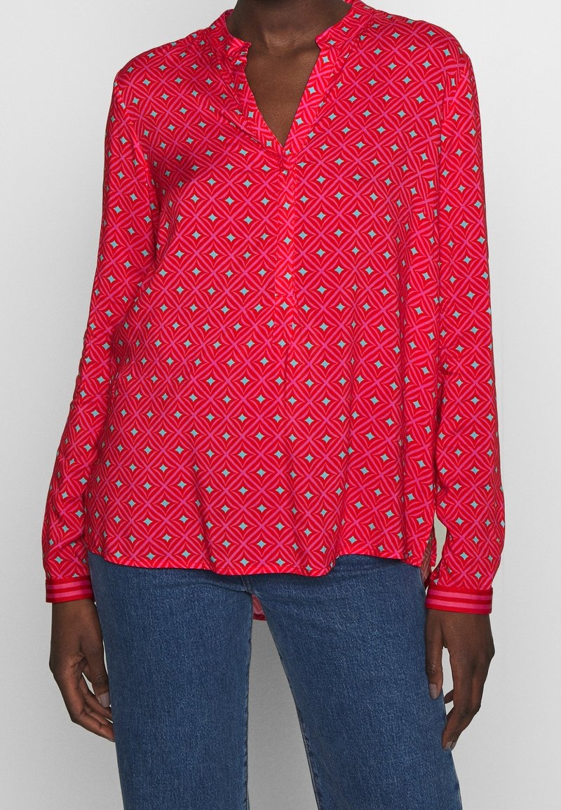 Blusa rossa a maniche lunghe con un motivo geometrico, dotata di colletto a scollo a V e bottoni, abbinata a jeans di jeans scuri.