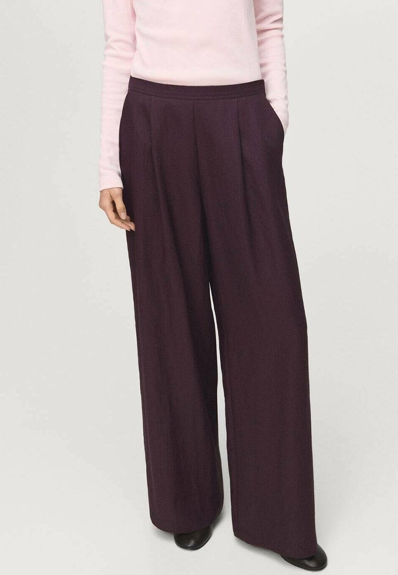 Femme portant un pantalon large violet foncé et un haut à manches longues rose clair, une main dans la poche et des chaussures noires visibles en dessous.