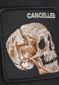 Parche de tela negro con un diseño de calavera bordado en beige y marrón, con la palabra "CANCELADO" en blanco encima de la calavera.