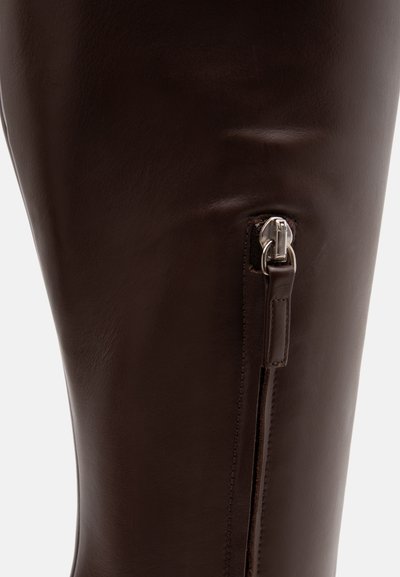 Gros plan d'une botte en cuir marron foncé montrant une fermeture éclair verticale argentée avec une languette en cuir sur le côté.