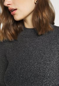 Femme aux cheveux bruns mi-longs portant un pull à col roulé côtelé noir scintillant et une boucle d’oreille créole en or.