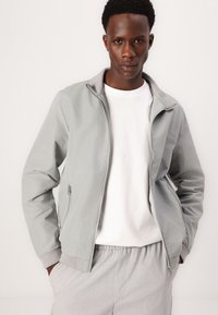 Helle graue Zip-Jacke mit Stehkragen, die eine glatte Textur und seitliche Taschen aufweist, über einem weißen T-Shirt und hellgrauen Hosen getragen.