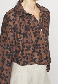 Blusa marrone con motivo floreale nero, dotata di colletto, chiusura con bottoni e una trama di tessuto fluida. Abbinata a pantaloni di colore chiaro.