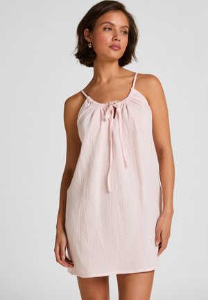 Junge Frau mit kurzen braunen Haaren, die ein blassrosa ärmelloses Kleid mit gerafftem Ausschnitt und gebundenem Schleifenband trägt, vor schlichtem Hintergrund stehend.