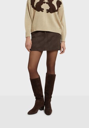 Frau trägt einen beigen Pullover mit dunklen Tierprints, einen dunkelbraunen Cord-Minirock, transparente Strumpfhosen und kniehohe dunkelbraune Wildlederstiefel.