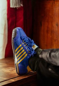 Baskets en velours bleu avec des rayures jaunes, semelle en caoutchouc et finition texturée. Associées à des chaussettes vert foncé, posées sur une surface en bois.