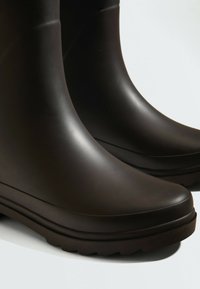 Paire de bottes de pluie en caoutchouc lisses marron foncé avec des semelles épaisses à crampons sur un fond clair.