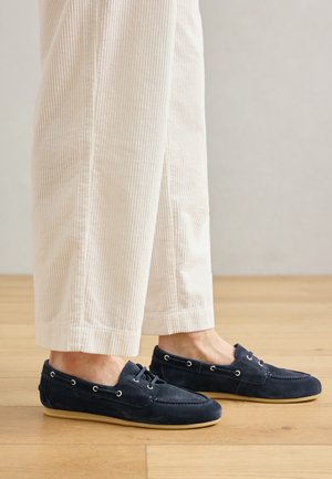 Chaussures bateau en suède bleu marine avec des œillets blancs contrastants, un lacet décoratif et une semelle en caoutchouc crème. Portées avec un pantalon en velours côtelé crème.