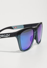 Ochelari de soare negri și teal, cu lentile mari și reflectante. Rama are un design îndrăzneț, unghiular, cu o finisare mată și un branding proeminent pe lateral.