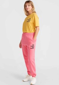 Gul bomullströja med svart text, kombinerad med rosa sweatpants med liknande svart text, båda i avslappnad passform och med muddar vid anklarna.