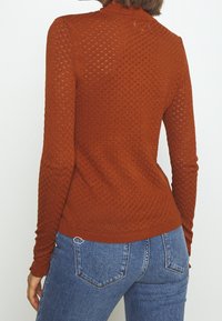 Pull tricoté couleur rouille avec un motif texturé, manches longues et col montant, associé à un jean en denim bleu.