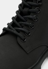 Gros plan d'une botte en cuir noir avec de gros lacets noirs enfilés à travers six paires d'œillets métalliques.