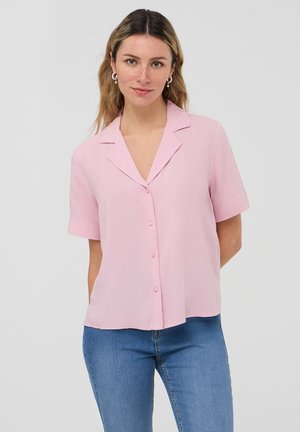 Blusa rosa a maniche corte con colletto a punta, chiusura a bottoni e tessuto lucido. Indossata con jeans in denim blu, dal taglio casual.
