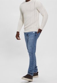 Witte kabelgebreide trui, blauwe slim-fit jeans en donkere sneakers. De trui heeft textuurpatronen; de jeans hebben een lichte slijtage.