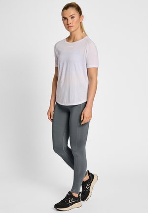 Kobieta stoi, ubrana w jasnofioletowy krótki rękaw, ciemnoszare legginsy i czarne buty do biegania na jednolitym białym tle.