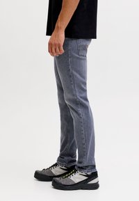 Mand stående i profil iført sort skjorte, falmede blå jeans og grå-sorte vandresko med sorte snørebånd på hvid baggrund.