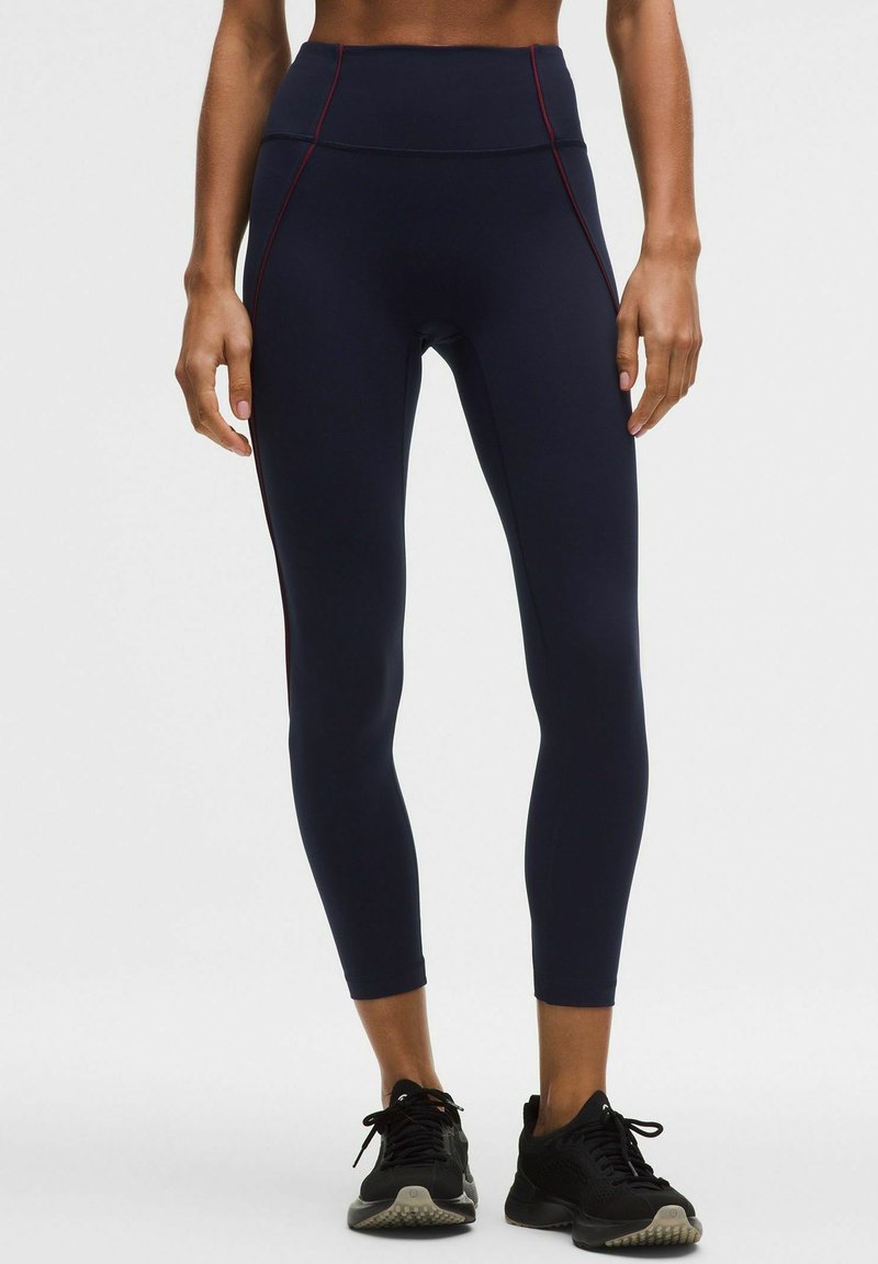 Žena oblečená v námornícky modrých športových legínách s vysokým pásom a červeným prešitím, s čiernymi bežeckými topánkami, stojaca na bielom pozadí.