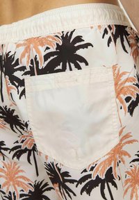 Pantaloni da bagno bianchi con stampa di palme nere e arancioni. Presentano una cintura elastica e una tasca quadrata sul retro. Texture liscia.