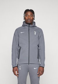 Grå zip-up hættetrøje lavet af blødt stof, med en kengurulomme, sorte lynlåse og en hvid logo på brystet og ærmet.