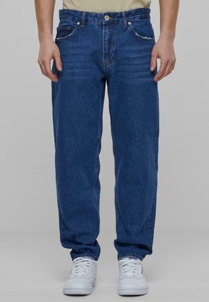 Jeans in denim blu con vestibilità dritta, cinque tasche e dettagli di sbiaditura leggera. Indossati con sneakers bianche, caratterizzate da un tessuto testurizzato.