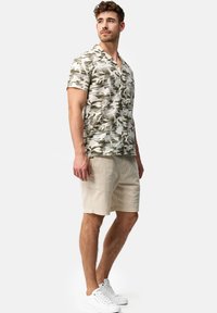 Chemise à manches courtes, à boutons, avec un motif de feuilles vertes et beiges, associée à un short beige et des baskets blanches. Design de col simple.